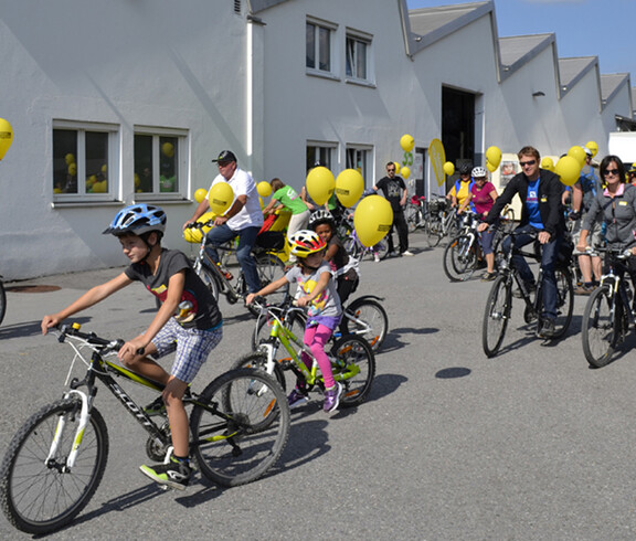 Ca. 20 Menschen mit an ihren Rädern gelb befestigten Luftballonen befinden sich auf einem großen asphaltierten Platz. 