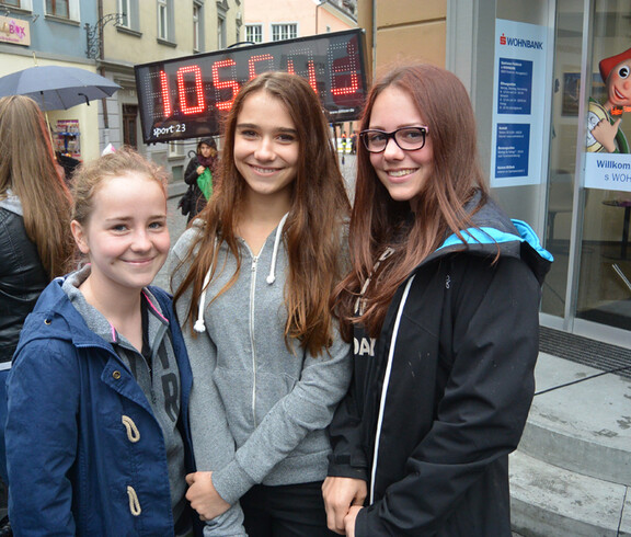 Drei Jugendliche mit jeweils langen braunen bzw. bonden Haaren stehen in der feldkircher Innenstadt für ein Foto zusammen. Sie tragen unter anderem graue, schwarze und blaue Kleidung. Sie wirken alle fröhlich und entspannt. 