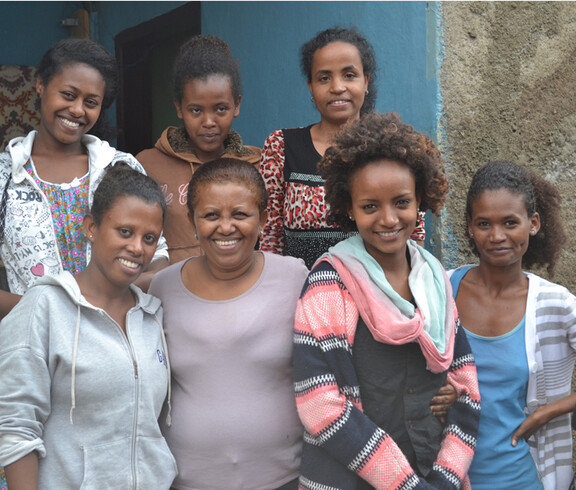 Junge und ältere Frauen posieren vor Hanna Orphans Home. Sie sind erfreut und lachen in die Kamera. Alle tragen ihre Haare schwarz. 