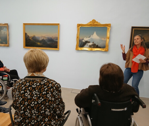 Gruppe von Bewohner*innen sitzt vor Gemälden im Museum, während eine Vermittlungsperson Kunstwerke erklärt