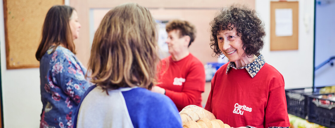 Eine freiwillige Mitarbeiterin der Caritas spricht lächelnd mit zwei Personen und verteilt Lebensmittel in einer Einrichtung.