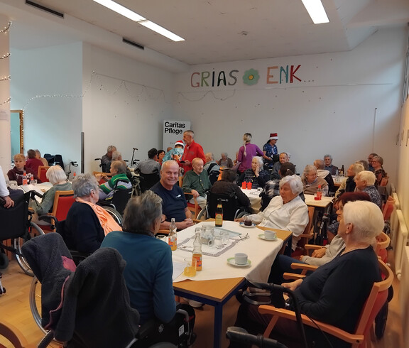 Viele Bewohner*innen sitzen an Tischen im Saal mit Weihnachtsdekoration und Getränken.