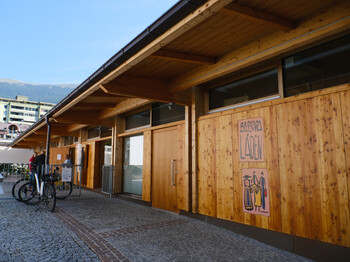 Zwei Personen stehen vor einem mit Holz verkleideten Gebäude und unterhalten sich.