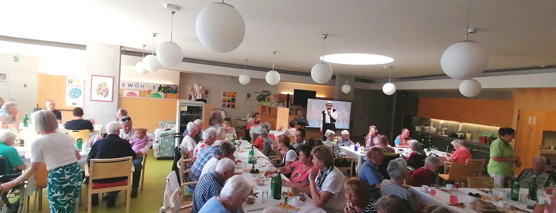 Beim Sommerfest im Pflegewohnhaus haben sich zahlreiche Bewohnerinnen und Bewohner, Angehörige sowie Mitarbeitende in einem festlich geschmückten Gemeinschaftsraum versammelt, genießen Speisen und Getränke an langen Tischen und verfolgen gemeinsam das Unterhaltungsprogramm.