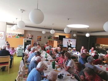 Beim Sommerfest im Pflegewohnhaus haben sich zahlreiche Bewohnerinnen und Bewohner, Angehörige sowie Mitarbeitende in einem festlich geschmückten Gemeinschaftsraum versammelt, genießen Speisen und Getränke an langen Tischen und verfolgen gemeinsam das Unterhaltungsprogramm.