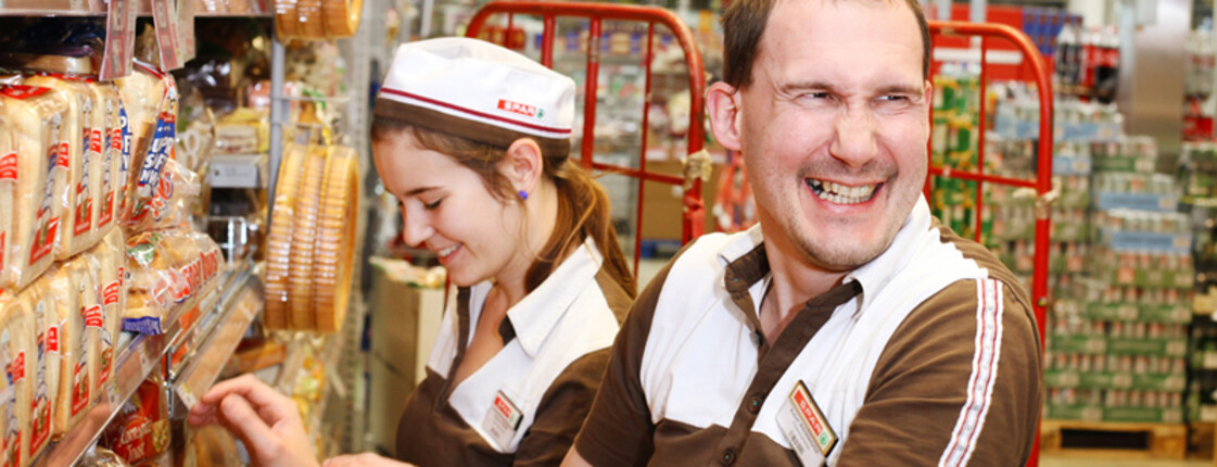 Zwei lachende SPAR-Mitarbeiter räumen Lebensmittel in ein Regal ein. 