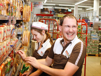 Zwei lachende SPAR-Mitarbeiter räumen Lebensmittel in ein Regal ein. 