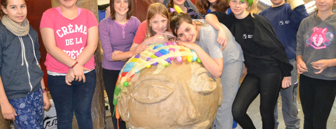 Jugendliche und eine Erwachsene Frau stellen sich um einen selbstgebastelten überdemensionierten Kopf. Sie sind in einem Raum und tragen Alltagskleidung. 