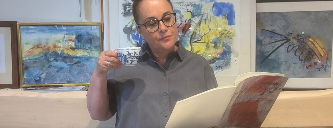Frau mit Brille hält eine Kaffeetasse und ein Kunstbuch in den Händen.