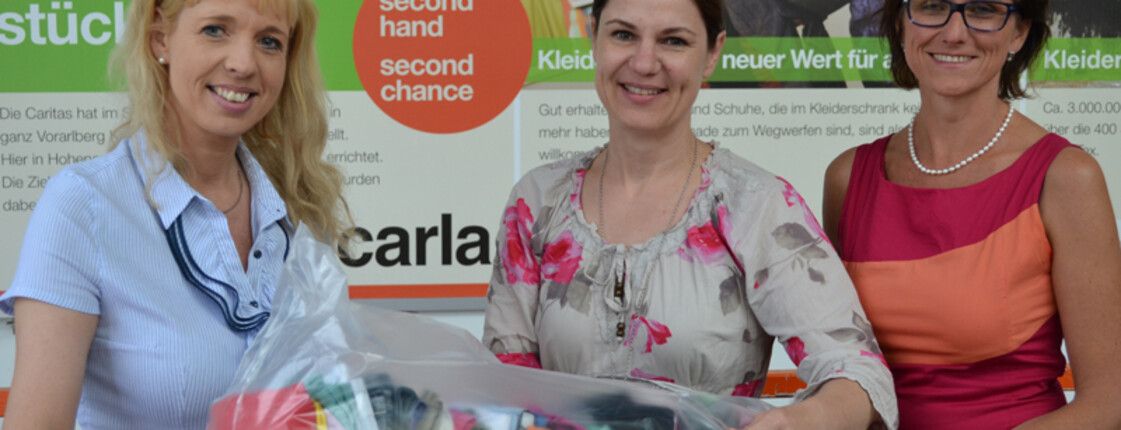 Drei Frauen mittleren Alters tragen einen Sack voll Kleider in der Hand. Im Hintergrund ist ein großes Plakat der Carla.