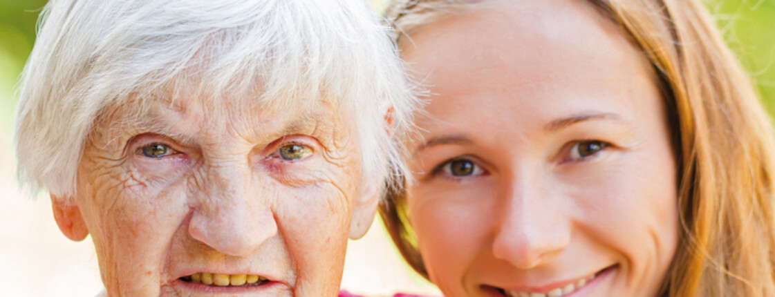 Ein junges Mädchen steht neben einer alten Frau. Die Frau hat graue Haare. Das Mädchen hat braune Haare und lächelt.