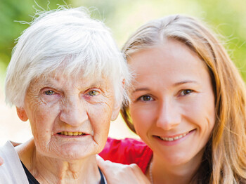 Ein junges Mädchen steht neben einer alten Frau. Die Frau hat graue Haare. Das Mädchen hat braune Haare und lächelt.