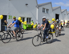Ca. 10 Menschen mit an ihren Rädern gelb befestigten Luftballonen befinden sich auf einem asphaltierten Platz. 