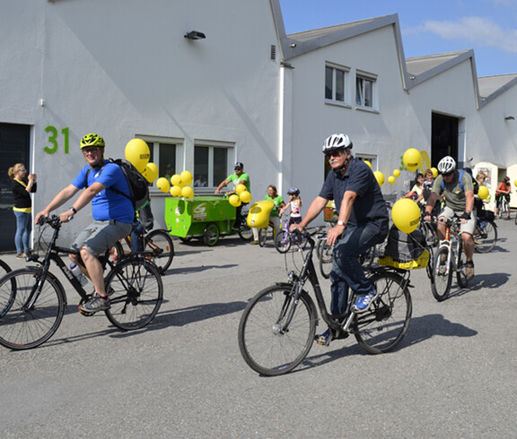 Ca. 10 Menschen mit an ihren Rädern gelb befestigten Luftballonen befinden sich auf einem asphaltierten Platz. 