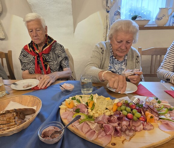 Mehrere Bewohnerinnen und Bewohner sitzen an einem gedeckten Holztisch und bedienen sich von einer großen, dekorativ angerichteten Jausenplatte mit verschiedenen Käsesorten, Schinken, Wurst, Trauben, Paprika und hartgekochten Eiern, während daneben Brotkorb, Aufstriche und Gläser mit Getränken stehen.
