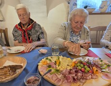 Mehrere Bewohnerinnen und Bewohner sitzen an einem gedeckten Holztisch und bedienen sich von einer großen, dekorativ angerichteten Jausenplatte mit verschiedenen Käsesorten, Schinken, Wurst, Trauben, Paprika und hartgekochten Eiern, während daneben Brotkorb, Aufstriche und Gläser mit Getränken stehen.