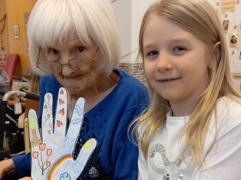Im Pflegewohnhaus steht ein Kind neben einer Bewohnerin im Rollstuhl und präsentiert gemeinsam ein liebevoll gestaltetes Bastelwerk im hellen Gemeinschaftsraum.