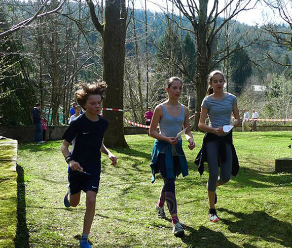 Zwei Mädchen und ein Junge laufen auf einer Wiese. Sie tragen eine Laufnummer in der Hand und tragen Sportbekleidung.