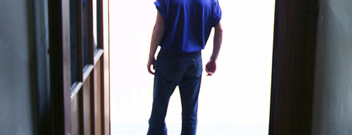 Ein männlicher Asylwerber wartet im Bereich des Eingangs seines Quartiers. Er hat schwarze Haare, trägt eine Jeans und ein blaues t-Shirt. Sein Blick richtet sich nach draußen. 