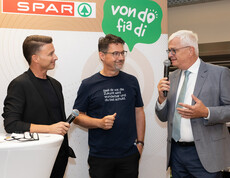 Zwei Personen führen Gespräch mit Mikrofon vor SPAR-Hintergrund im Markt zwischen Regalen.