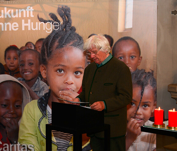 In der Pfarrkirche von Frastanz steht ein Mann fortgeschrittenen Alters mit weißen Haaren am Rednerpult. Der Hintergrund ziert ein großes Plakat mit afrikanischen Kindern. 