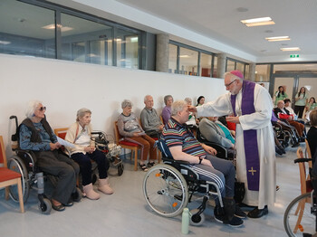 Priester zeichnet einer Person im Rollstuhl das Aschenkreuz im Gemeinschaftsraum.