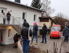 Neues Haus von außen, mit mehreren Menschen und einem roten Auto.