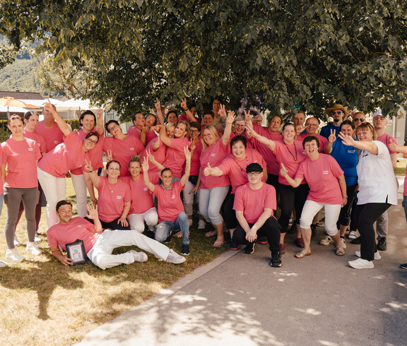 Große Gruppe von Mitarbeitenden in pinken Shirts posiert im Garten unter einem Baum beim Sommerfest