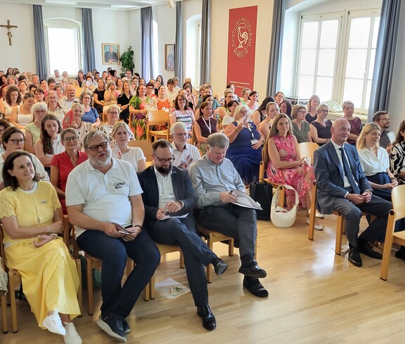 Viele Leute sitzen im Publikum auf Sesselreihen nebeneinander und schauen in eine Richtung