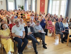 Viele Leute sitzen im Publikum auf Sesselreihen nebeneinander und schauen in eine Richtung