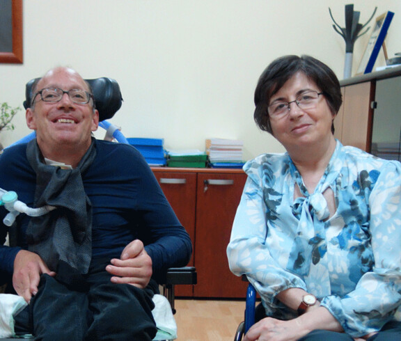 Ein lächelnder Mann im Rollstuhl sitzt neben einer Frau in einem Büro – beide wirken freundlich und offen.