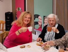 Zwei Personen sitzen am Tisch mit Getränken und Gebäck, bunte Banner im Hintergrund