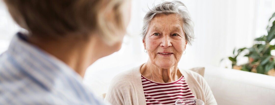 Eine Seniorin sitzt auf einem Sofa und bekommt von einer anderen Frau ein Glas Wasser gereicht.