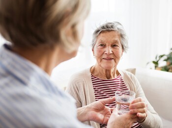 Eine Seniorin sitzt auf einem Sofa und bekommt von einer anderen Frau ein Glas Wasser gereicht.
