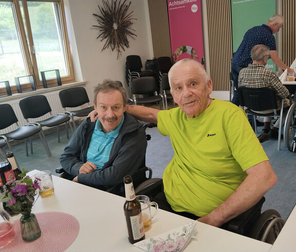 Zwei Bewohner sitzen bei Getränken an einem Tisch, eine Person legt den Arm um die andere während der Muttertagsfeier.