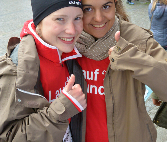 Zwei junge Frauen nehmen an einem Laufwettbewerb in der feldkircher Innenstadt teil und stehen zu einem Foto zusammen. Sie haben beide braune Jacken und rote Oberteile an. 
