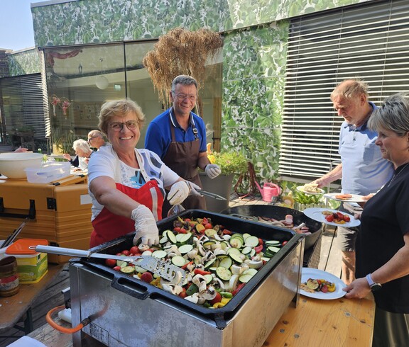 Beim Sommerfest im Pflegewohnhaus wird im Innenhof frisch gegrillt: Mehrere Personen bereiten an einer großen Grillplatte buntes Gemüse und Würstchen zu, während Gäste ihre Teller füllen und das gemeinsame Festessen genießen.
