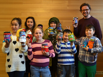 Bunt gekleidete Kleinkinder zeigen mit ihrem Lehrer im Lerncafé Lustenau das Ergebnis ihrer Bastelstunden. Alte Marmeladengläser wurden mit Filtz überzogen und dabei mit unterschliedlichen Farben desselben Gesichter kreiert. 