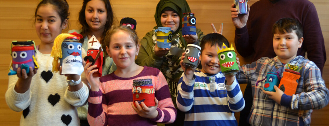 Bunt gekleidete Kleinkinder zeigen mit ihrem Lehrer im Lerncafé Lustenau das Ergebnis ihrer Bastelstunden. Alte Marmeladengläser wurden mit Filtz überzogen und dabei mit unterschliedlichen Farben desselben Gesichter kreiert. 