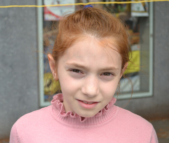 Ein junges Mädchen im Volksschulalter blickt in die Kamera. Sie hat rote Haare und trägt ein rosa Oberteil. Ihre Haare hat sie nach hinten zusammengebunden. 