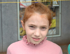 Ein junges Mädchen im Volksschulalter blickt in die Kamera. Sie hat rote Haare und trägt ein rosa Oberteil. Ihre Haare hat sie nach hinten zusammengebunden. 