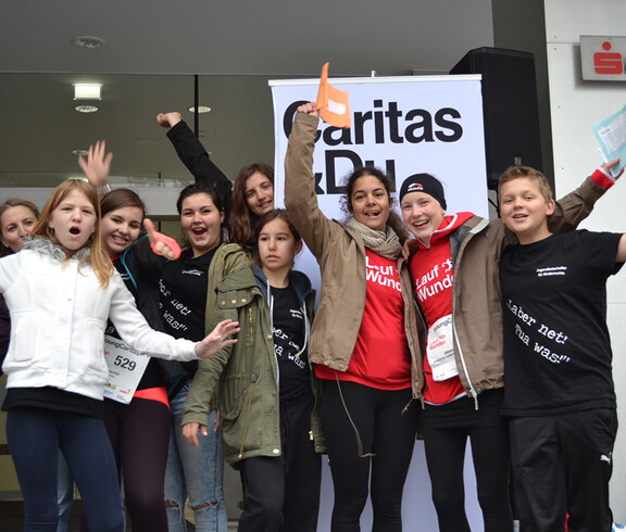 Kinder und Jugendliche stehen vor der Sparkasse in Feldkirch, jubeln und strecken ihre Hände in die Luft. Im Hintergrund befindet sich ein Caritas Roll-Up. Sie nahmen bei einem Laufwettbewerb teil. Manche tragen gemeinsame Trikots. Alle wirken freundlich und die Stimmung ist ausgelassen. 