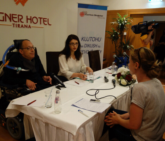 Ein Mann im Rollstuhl und zwei Frauen nehmen an einer Pressekonferenz von Caritas Albanien im Hotel Rogner in Tirana teil.