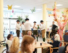 Bewohnerinnen und Bewohner eines Pflegewohnhauses erleben beim gemeinsamen Maibaumaufstellen musikalische Unterhaltung und geselliges Beisammensein im Aufenthaltsraum.