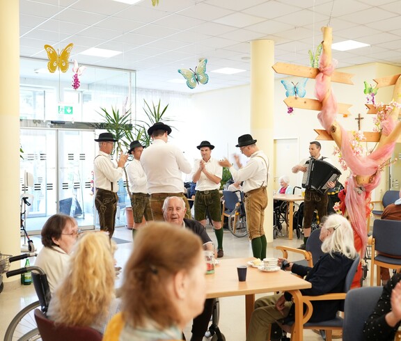 Bewohnerinnen und Bewohner eines Pflegewohnhauses erleben beim gemeinsamen Maibaumaufstellen musikalische Unterhaltung und geselliges Beisammensein im Aufenthaltsraum.