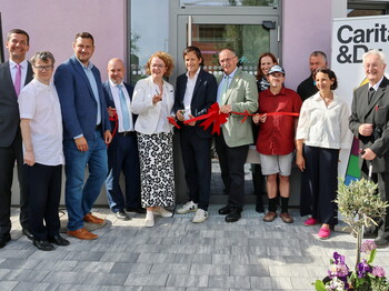 Gruppe von Menschen bei der Eröffnung vor einem Gebäudeeingang, mehrere Personen halten eine große rote Schleife und Scheren. Im Hintergrund ist ein Eingang mit Glasfront sowie ein Banner mit der Aufschrift „Caritas & Du“ zu sehen.