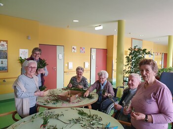 Bewohnerinnen und Bewohner des Pflegewohnhauses sitzen gemeinsam an mehreren Tischen und binden Palmbuschen. Zwischen frischen Zweigen, Bändern und Bastelmaterial entstehen in gemütlicher Runde kunstvolle Ostersträuße. Die fröhliche Aktivität findet im farbenfrohen Aufenthaltsbereich statt.