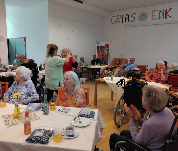 Raum mit vielen Bewohner*innen an Tischen, ein Paar tanzt, andere klatschen; Schild „Grias enk“ an der Wand sichtbar.