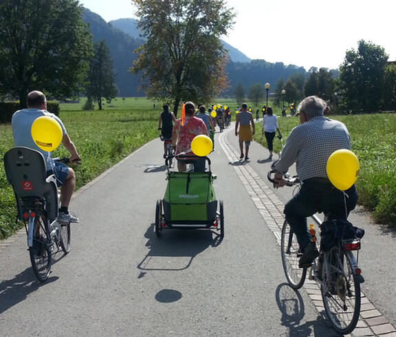 Ca. 10 sich im Bild befindende Personen fahren auf einer flachen Feldstraße Fahrrad. Dabei hat jeder einen gelben Luftballon am Fahrrad angebracht. 