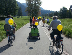 Ca. 10 sich im Bild befindende Personen fahren auf einer flachen Feldstraße Fahrrad. Dabei hat jeder einen gelben Luftballon am Fahrrad angebracht. 